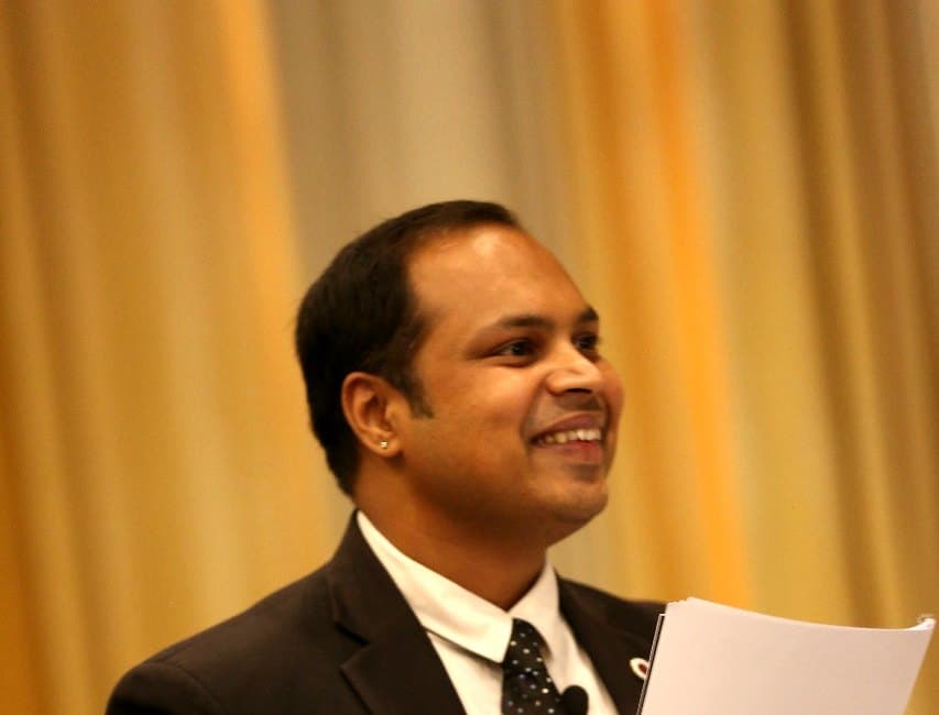 An advocate in a dark suit smiling while speaking, papers in hand