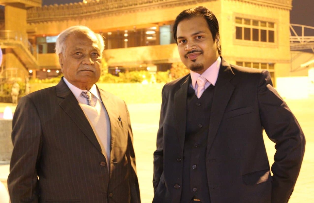 Adhiraj Surana standing beside his grandfather, Shri Devraj Singh Surana, both in dark suits, outdoors in the evening
