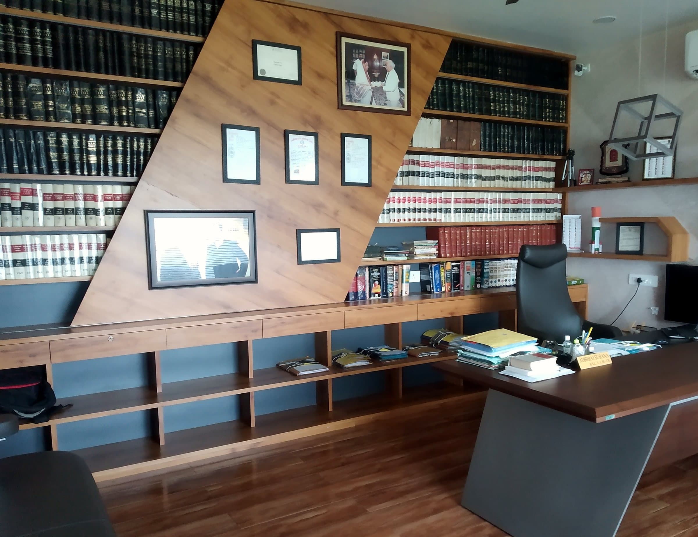 Chambers lined with bound law reports, framed certificates and photographs on a diagonal wooden panel, and a desk with case files